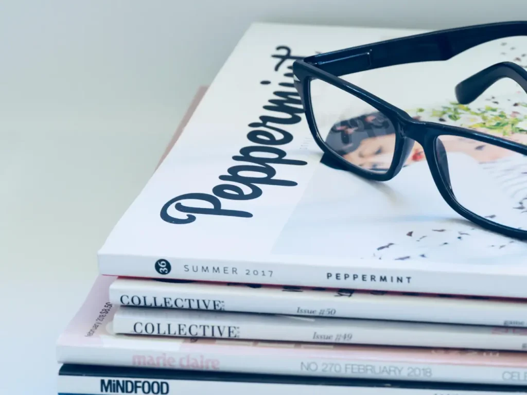 A minimalist lifestyle shot featuring a pair of black-rimmed glasses resting on a stack of various fashion and lifestyle magazines.