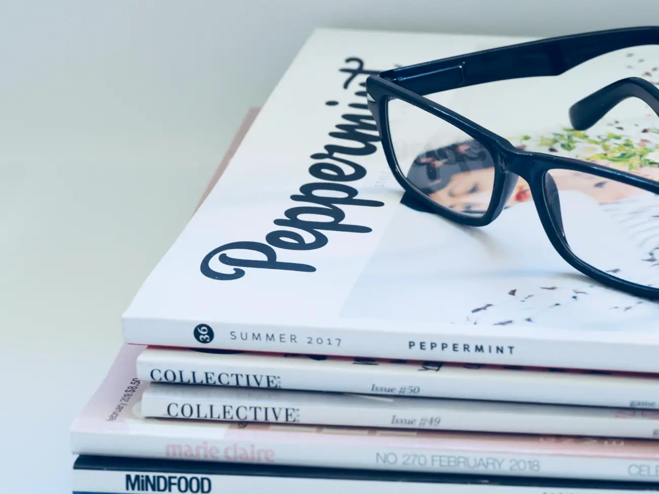 A minimalist lifestyle shot featuring a pair of black-rimmed glasses resting on a stack of various fashion and lifestyle magazines.