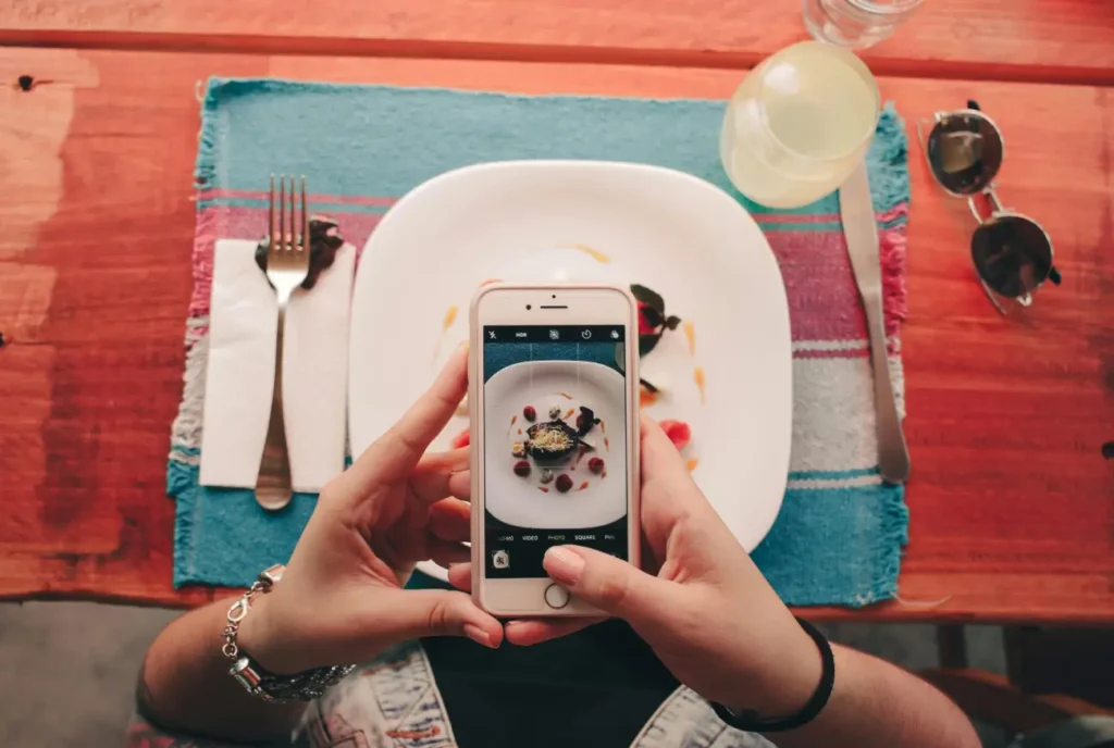 A top-down view of a person using a white smartphone to take a photo of a gourmet plated dish at a restaurant table.