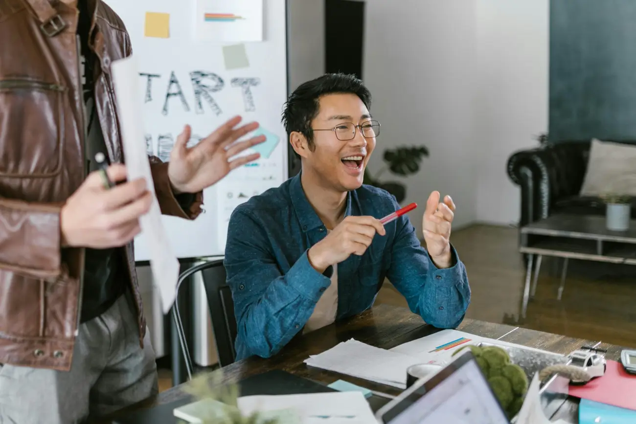 An office scene showing a man with glasses laughing and gesturing during a brainstorming session with a colleague.