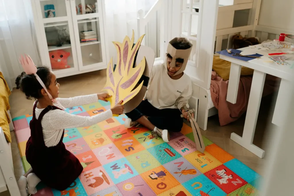 Two young children playing indoors on a colorful alphabet mat, dressed in homemade cardboard costumes.
