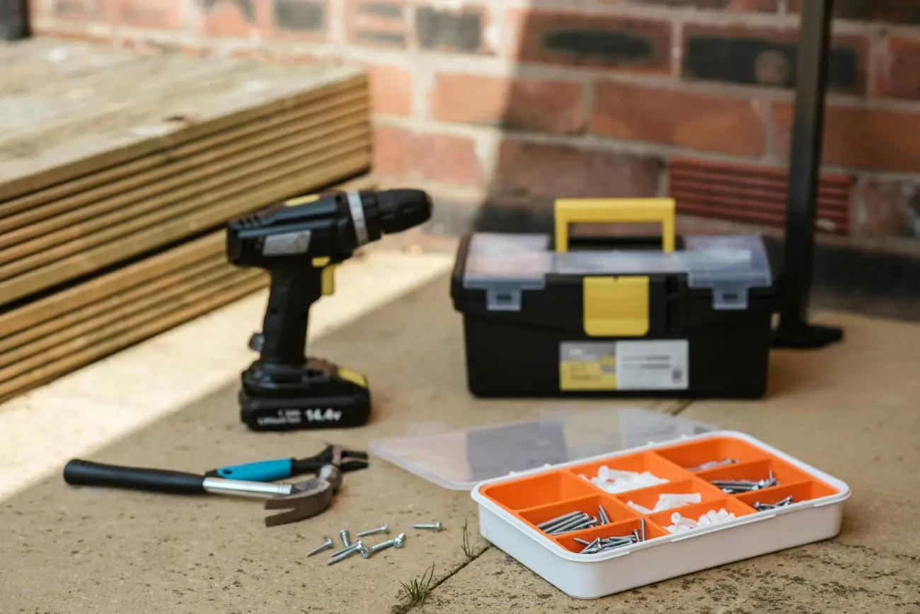 A collection of home improvement tools on a concrete floor, including a black power drill, hammer, and toolbox.