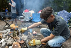 A man wearing protective gloves crouched by a campfire, carefully grilling skewers of meat in a forest setting.