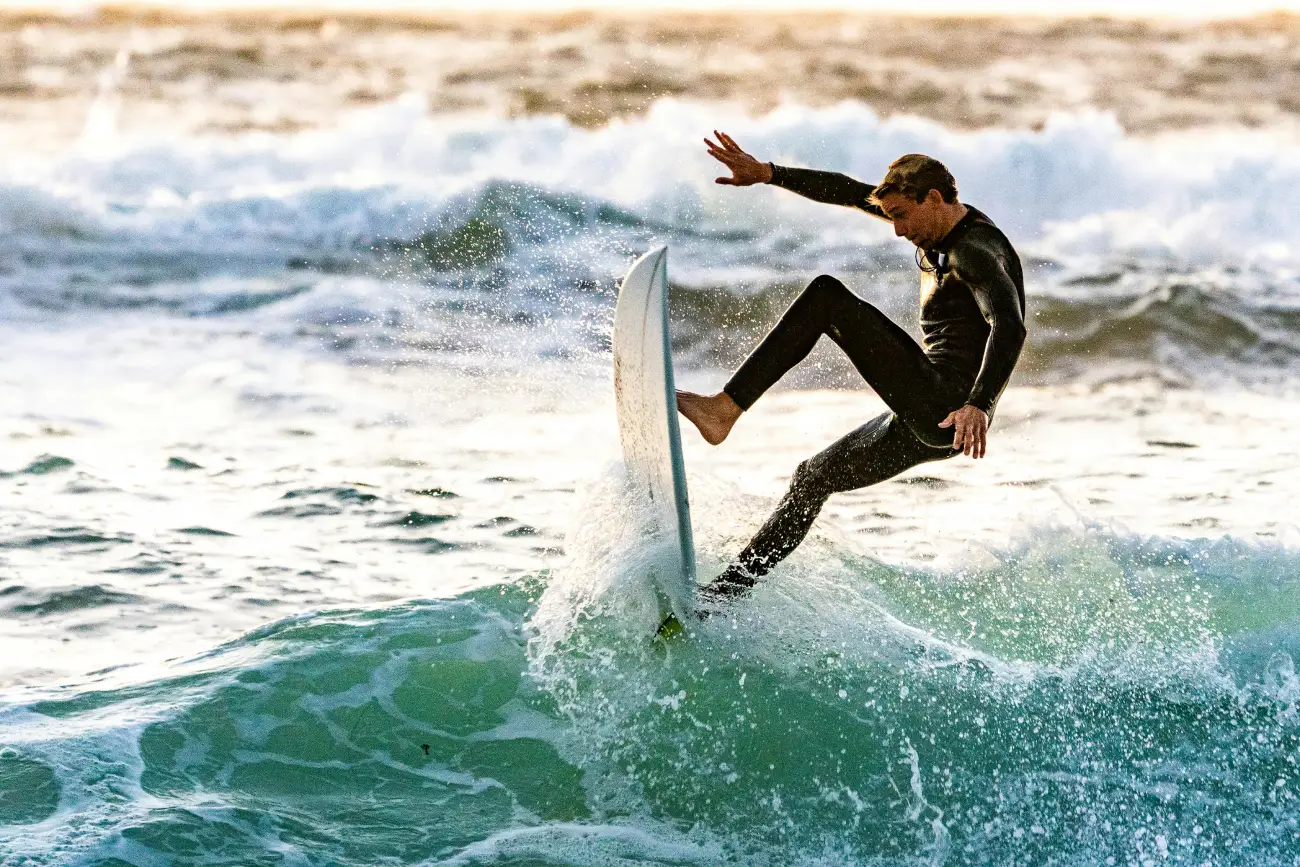 A high-action shot of a surfer in a black wetsuit catching air off the crest of a breaking ocean wave.